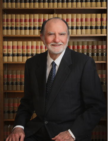 A man in a suit and tie stands in front of a bookshelf full of books.