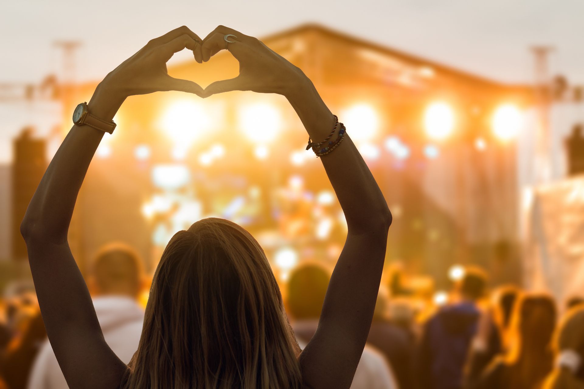 Person in crowd making heart with hands at outdoor concert stage.