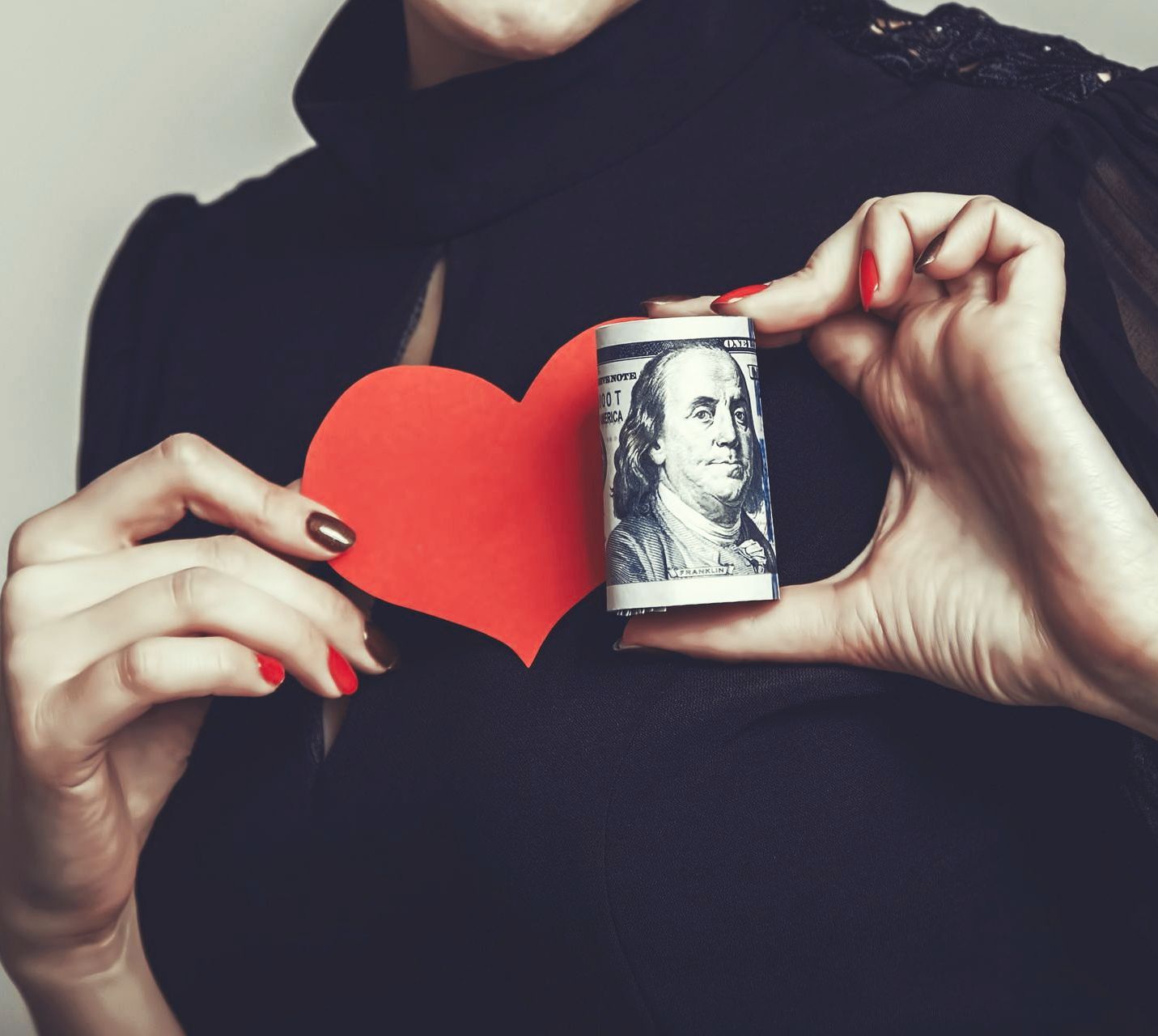 Woman holding a red heart and a roll of $100 bills against a black top.