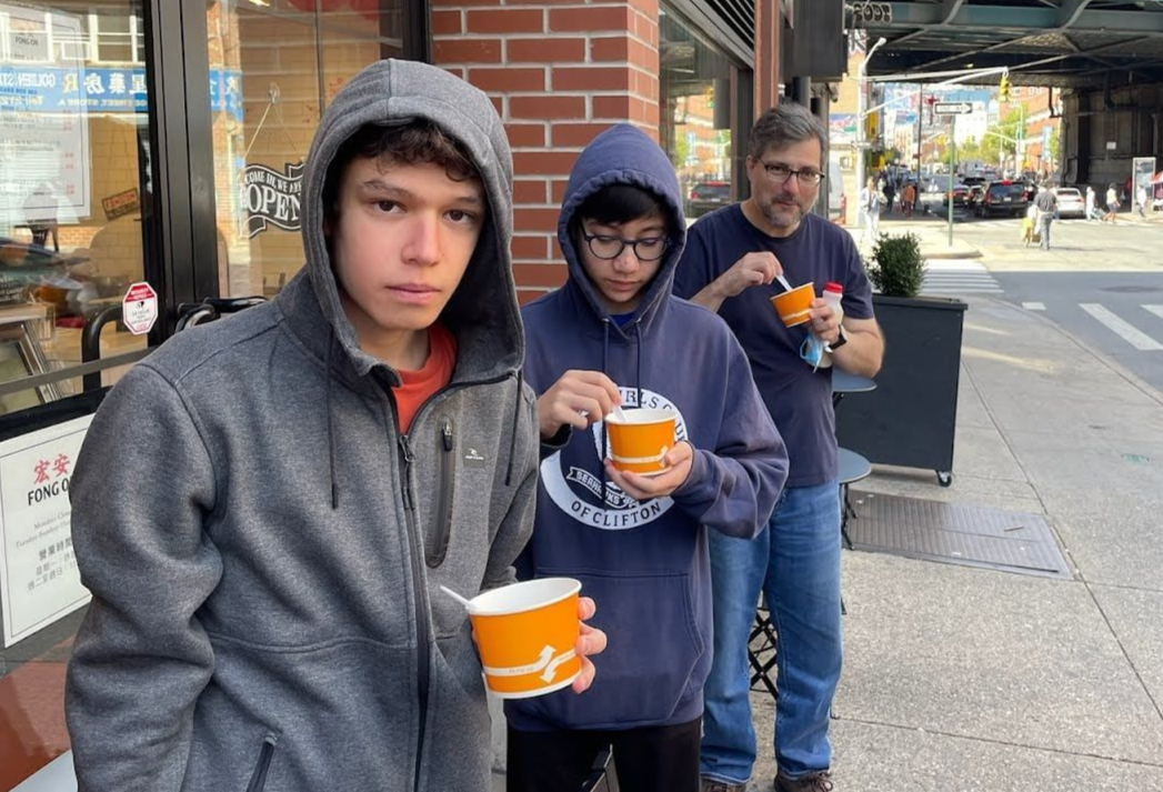 Three people in hoodies holding orange cups on a city sidewalk outside a brick building.