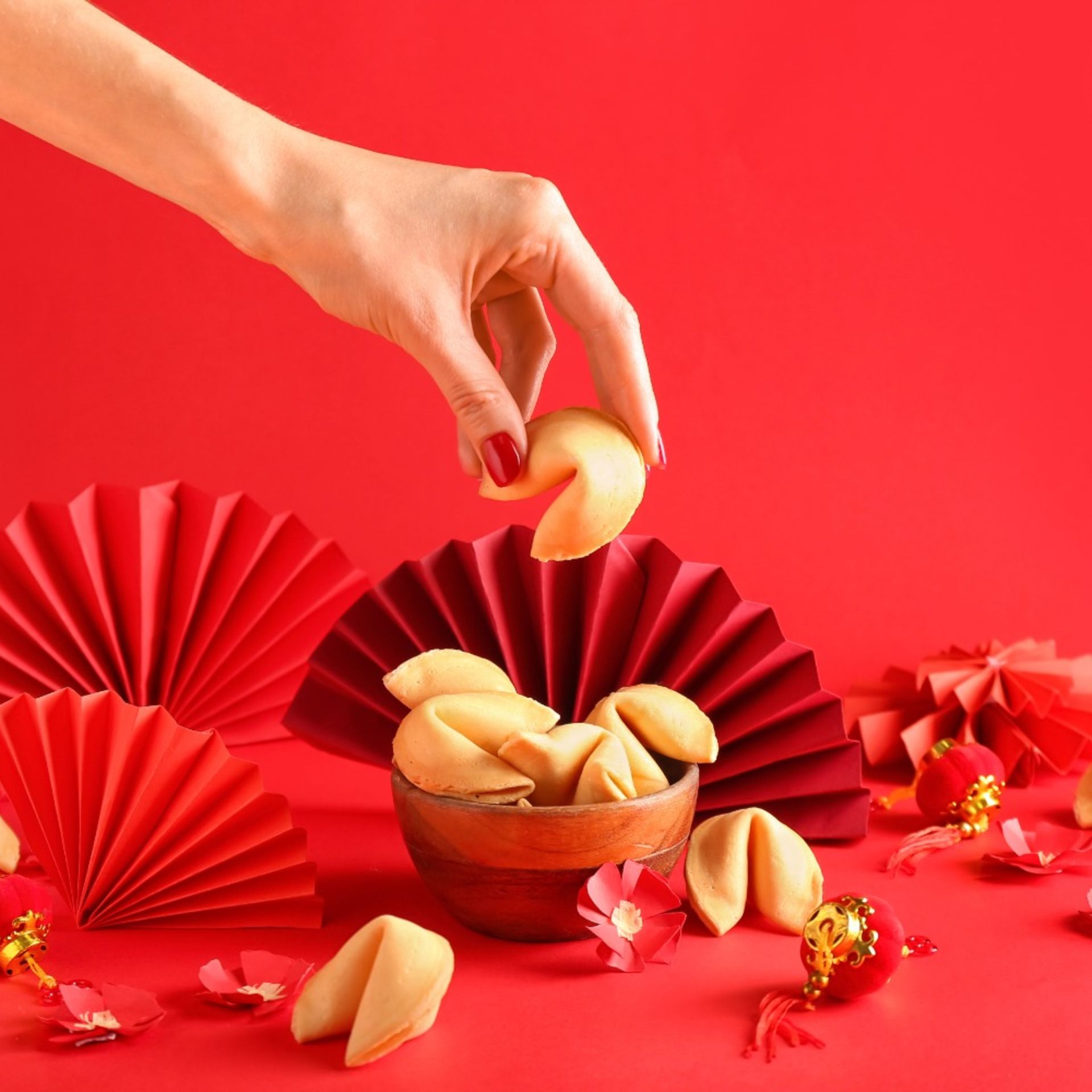 Hand placing a fortune cookie in a wooden bowl on a red festive background with folded paper fans