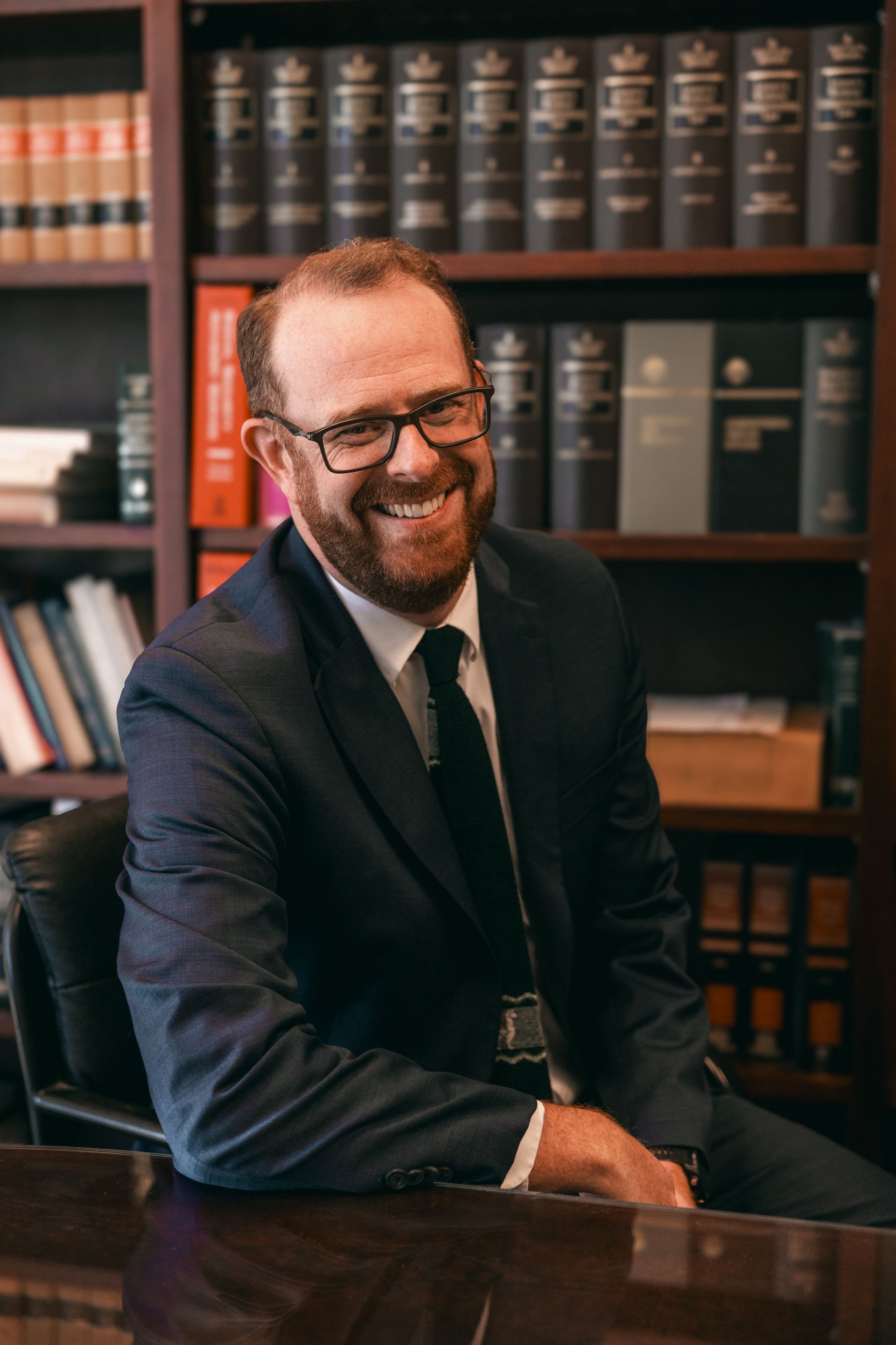 Man in suit smiling, seated in a wood-paneled office with bookshelves.