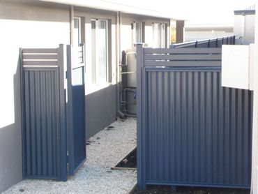Blue metal gates open onto a narrow path with a corrugated fence.