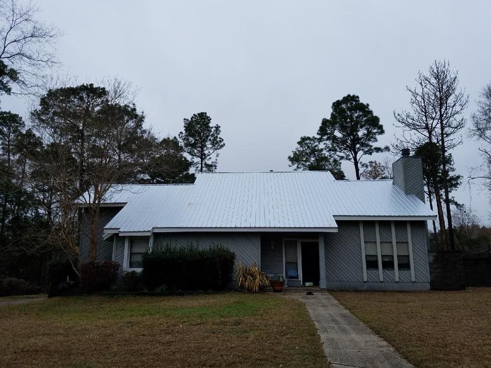 Two construction workers on a rooftop, one using a drill, both wearing safety vests and helmets. Gray house with metal roof on a cloudy day, surrounded by trees and a pathway leading to the door.