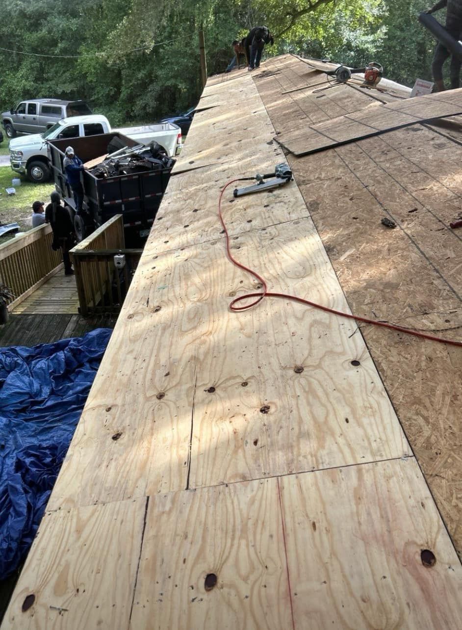 Roofers working on a house roof. Plywood sheets being installed. Trucks and debris visible. Roofers working on a house roof. Plywood sheets being installed. Trucks and debris visible.