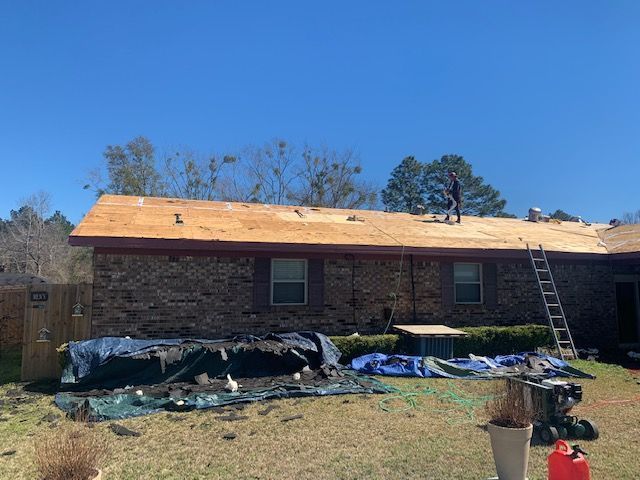 Roofing crew working on a house with exposed wooden roof. Tarps cover the yard and a ladder is leaning against the house.