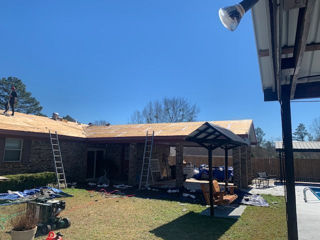 Workers on a roof, removing old shingles and replacing with new. Ladders, tools, and materials on the ground.