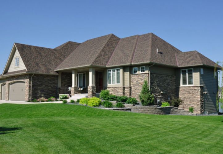 Large, modern house with brown roof and brick exterior, on a manicured lawn under a blue sky.