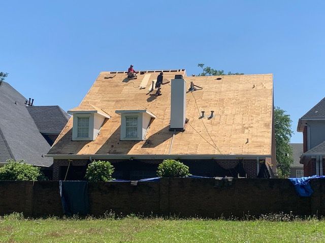 Workers on a rooftop, replacing shingles. Clear blue sky, wood roof, chimney, dormers.
