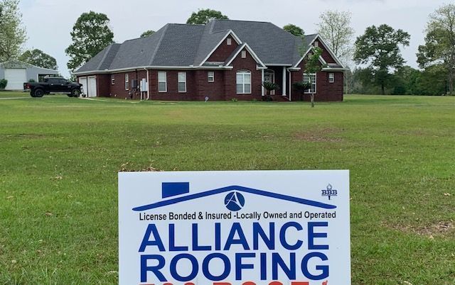 Sign for Alliance Roofing in front of a brick house with a dark roof and a truck in the yard.