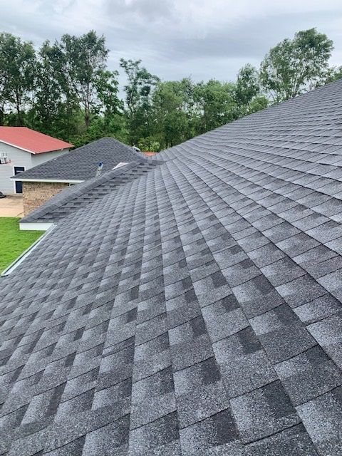 Gray asphalt shingle roof on a house, with green trees and cloudy sky in the background.
