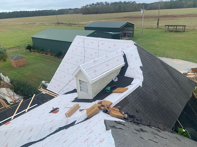Roof partially covered with white underlayment, and new shingles. Gable dormer attached. Green barn in the background.