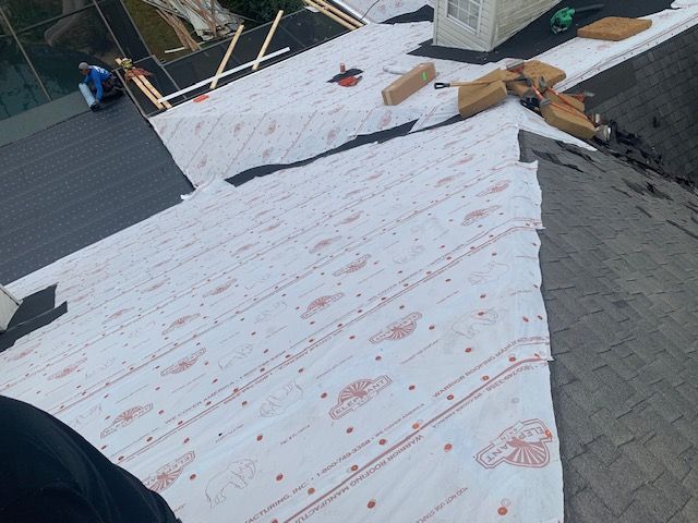 Roof partially covered with white underlayment, dark shingles, and a person working.