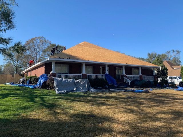 Workers repairing a roof on a brick home. Tarps cover the yard, a clear blue sky overhead.
