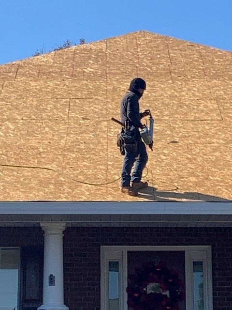 Roofer in dark clothing with a nail gun on a roof covered in wood sheeting. Bright blue sky.