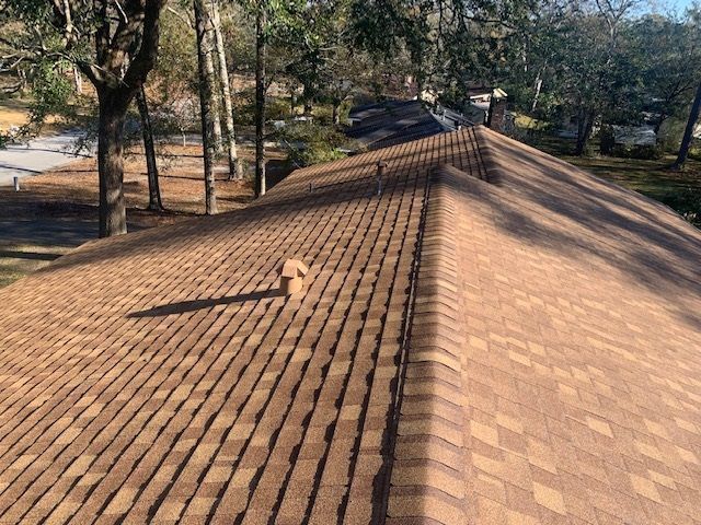 Brown shingled roof with a vent pipe, seen from above. Trees and a road are visible in the background.