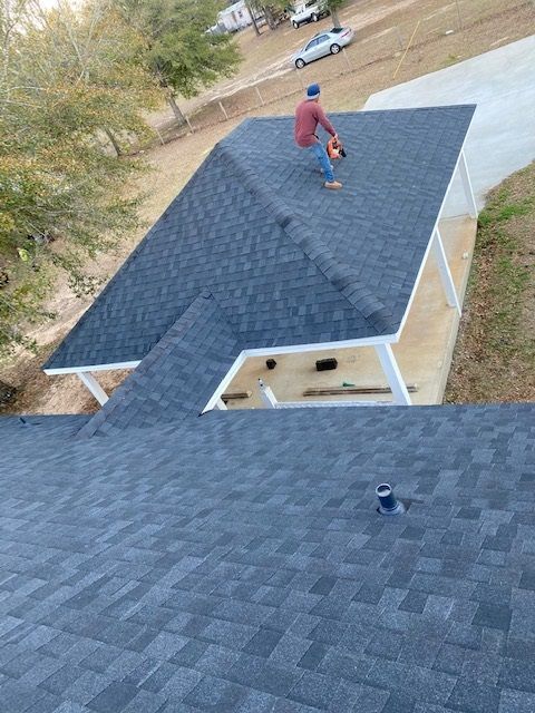 Roofer on a newly shingled roof, using a power tool. Another roof section in the foreground.