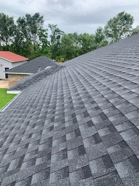 Dark gray shingle roof on a house, with a cloudy sky in the background and green trees.