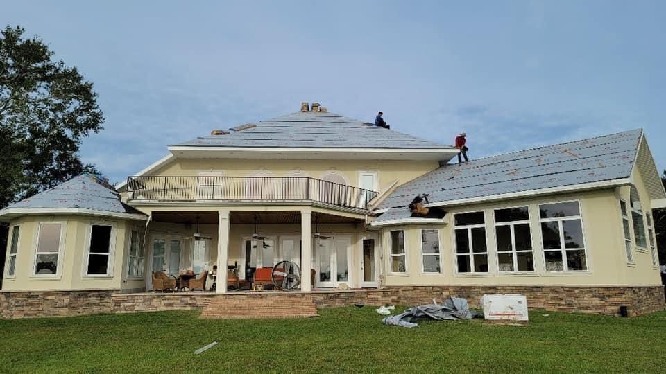 Workers on the roof of a two-story beige house with gray roofing, replacing shingles.