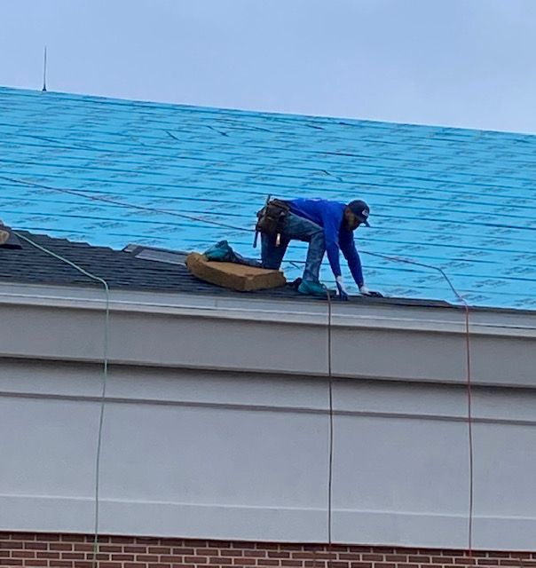 Roofer in blue shirt kneels on a roof, working. Blue tarp underlayment visible.