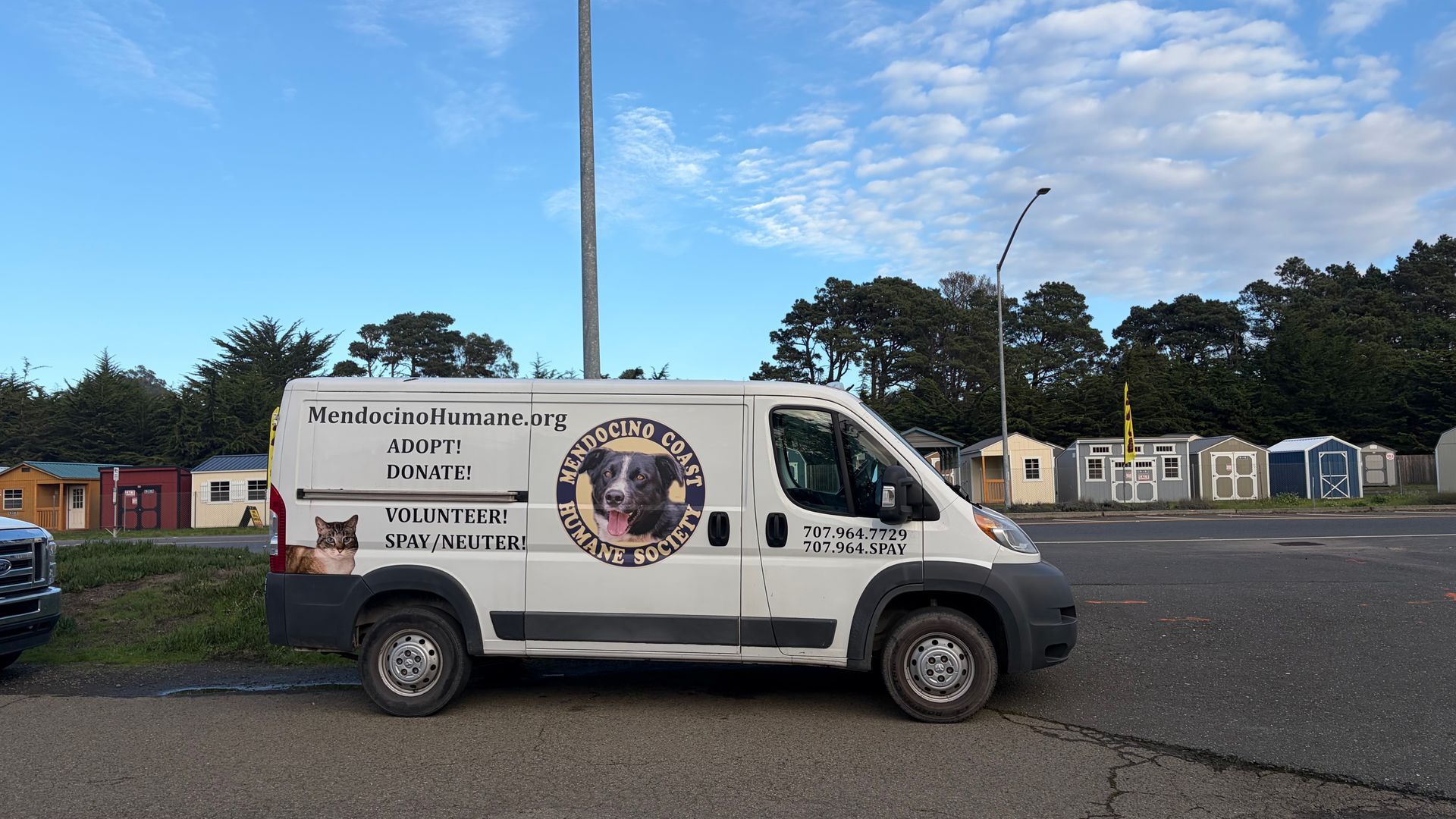 White van with dog logo parked in front of small shelters, under a blue sky.