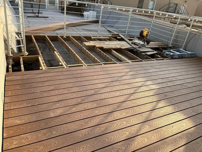 Wooden deck under construction with exposed joists, railing, and a worker using power tools.