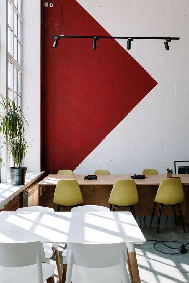 Modern cafe interior with red geometric wall art, wooden table, and green chairs by large windows