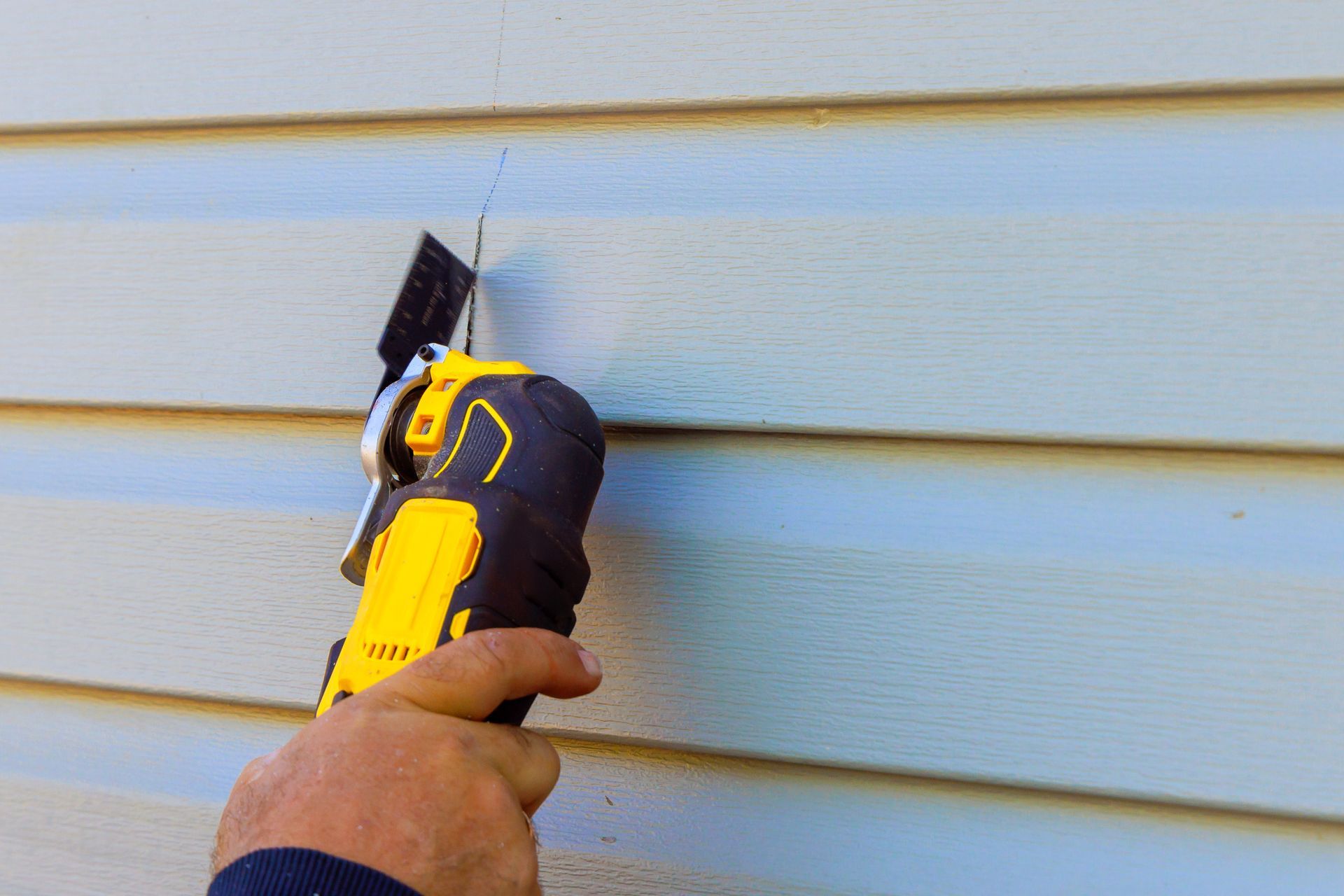 A siding replacement contractor cutting gray vinyl lap siding with a yellow oscillating power tool. A siding replacement contractor cutting gray vinyl lap siding with a yellow oscillating power tool.