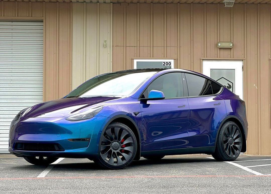 Blue and purple color-shifting Tesla Model Y parked in front of a building with a black roof and black wheels.