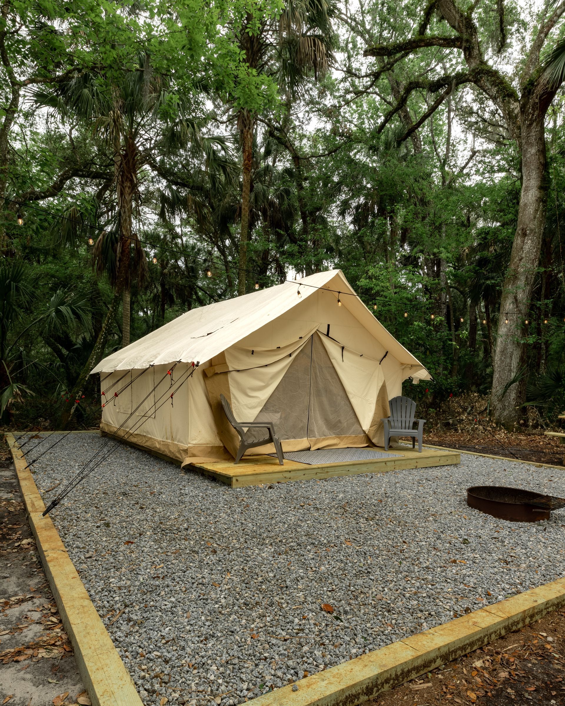 Tan canvas tent on gravel, set in a wooded campsite with a fire pit and trees.