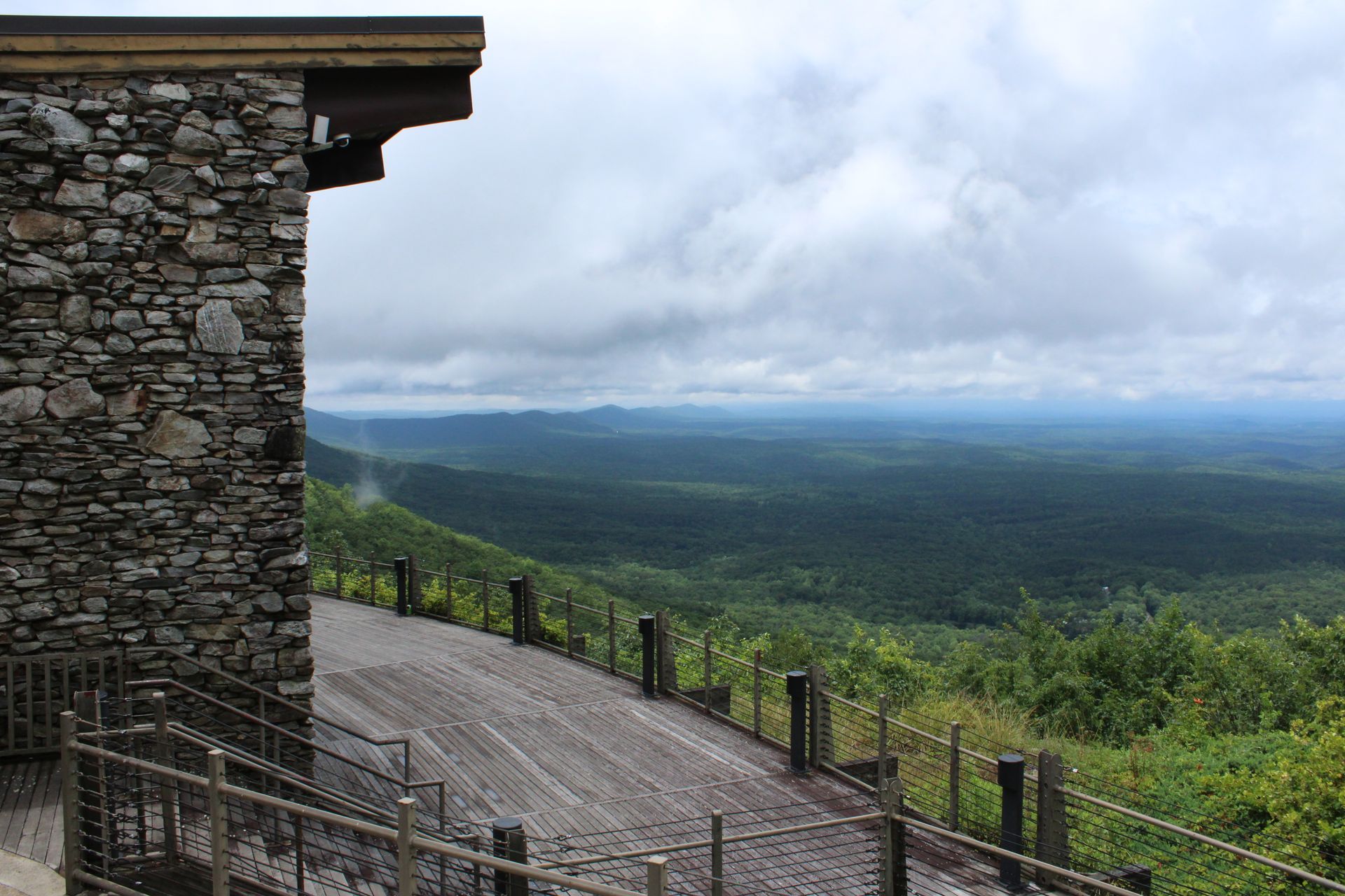 Stone building edge overlooking a vast green forest under a cloudy sky.