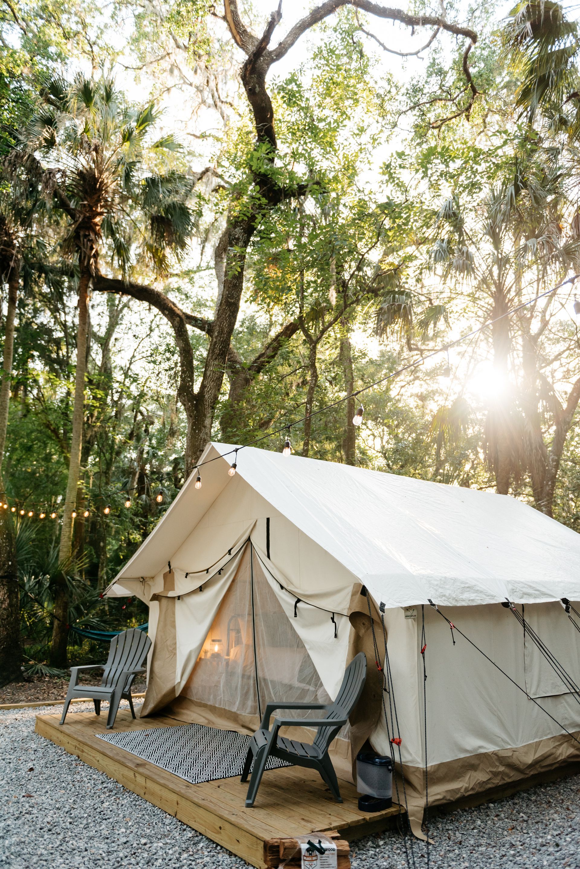 Canvas tent on a wooden platform, two chairs, string lights, forest background, sunny.