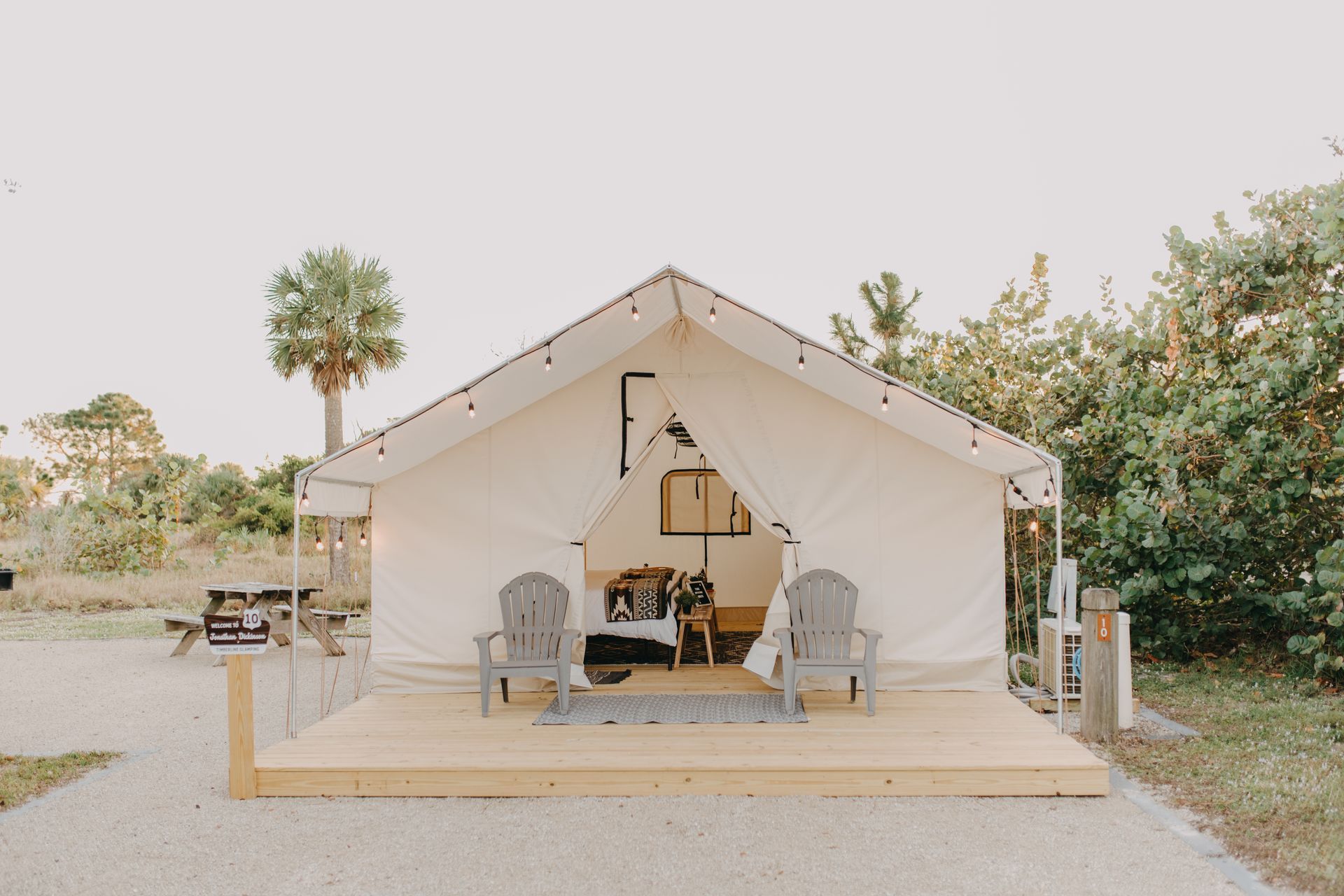 White canvas glamping tent on a wooden platform with two chairs, surrounded by gravel and foliage.