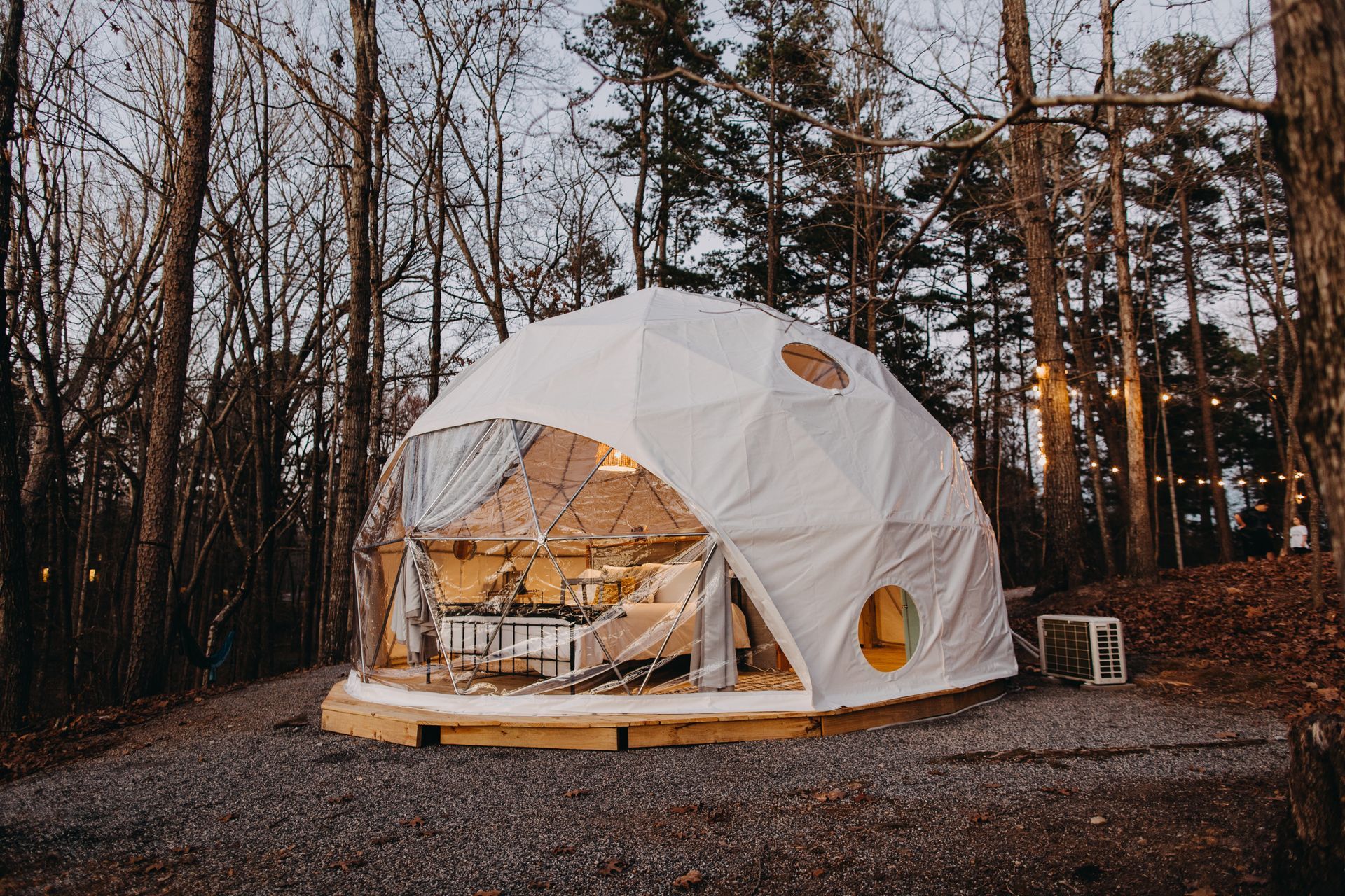 A white geodesic dome tent sits on gravel, with a partially transparent front, in a forest at dusk.