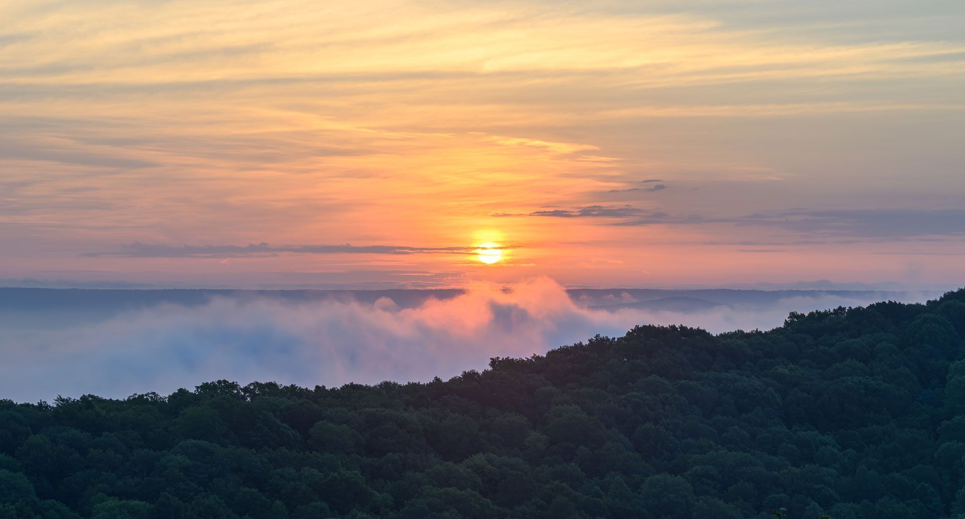 Sunset over a forested mountain with low-lying clouds and colorful sky.