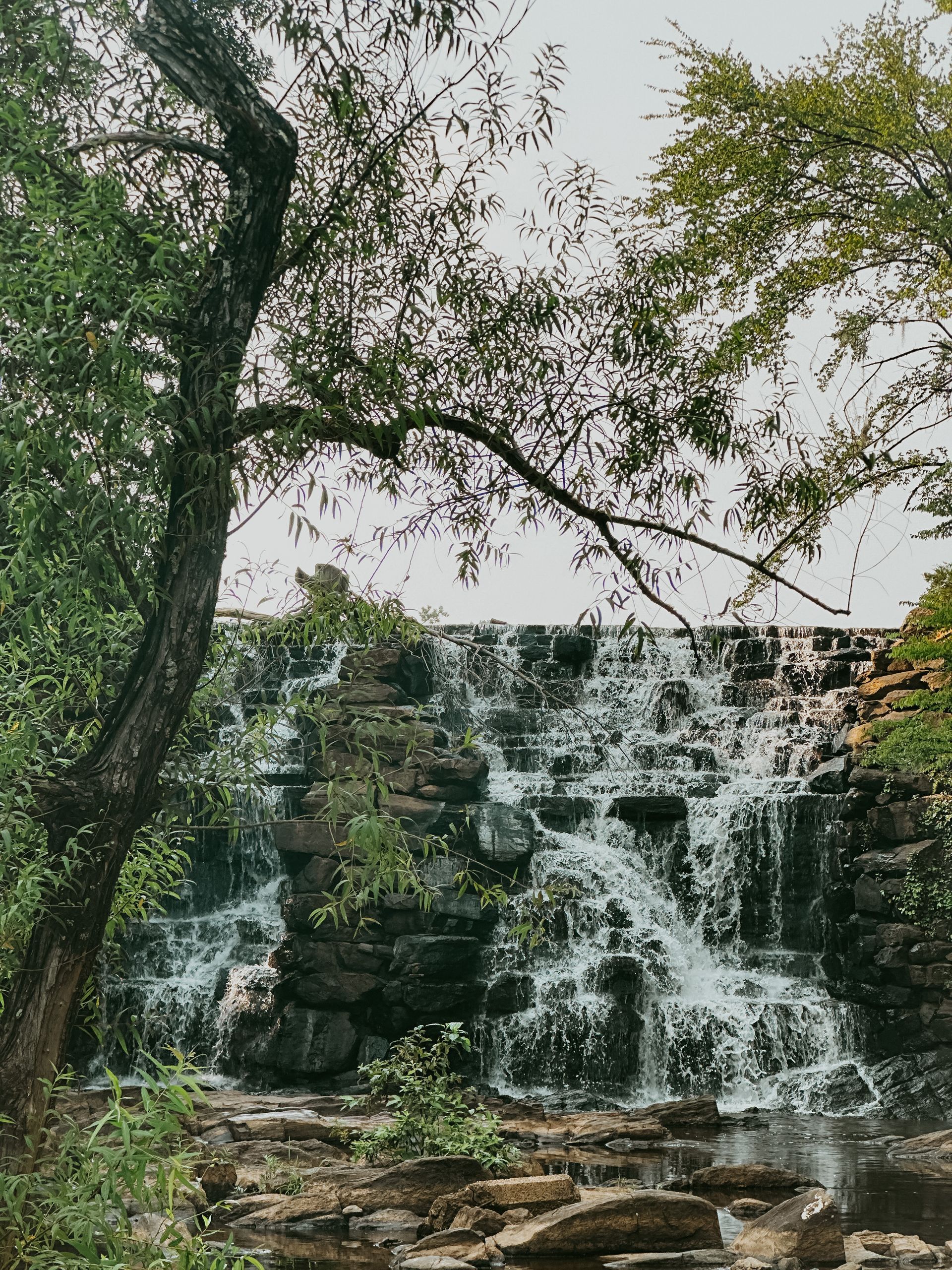 Waterfall cascading over stone steps, surrounded by green trees.