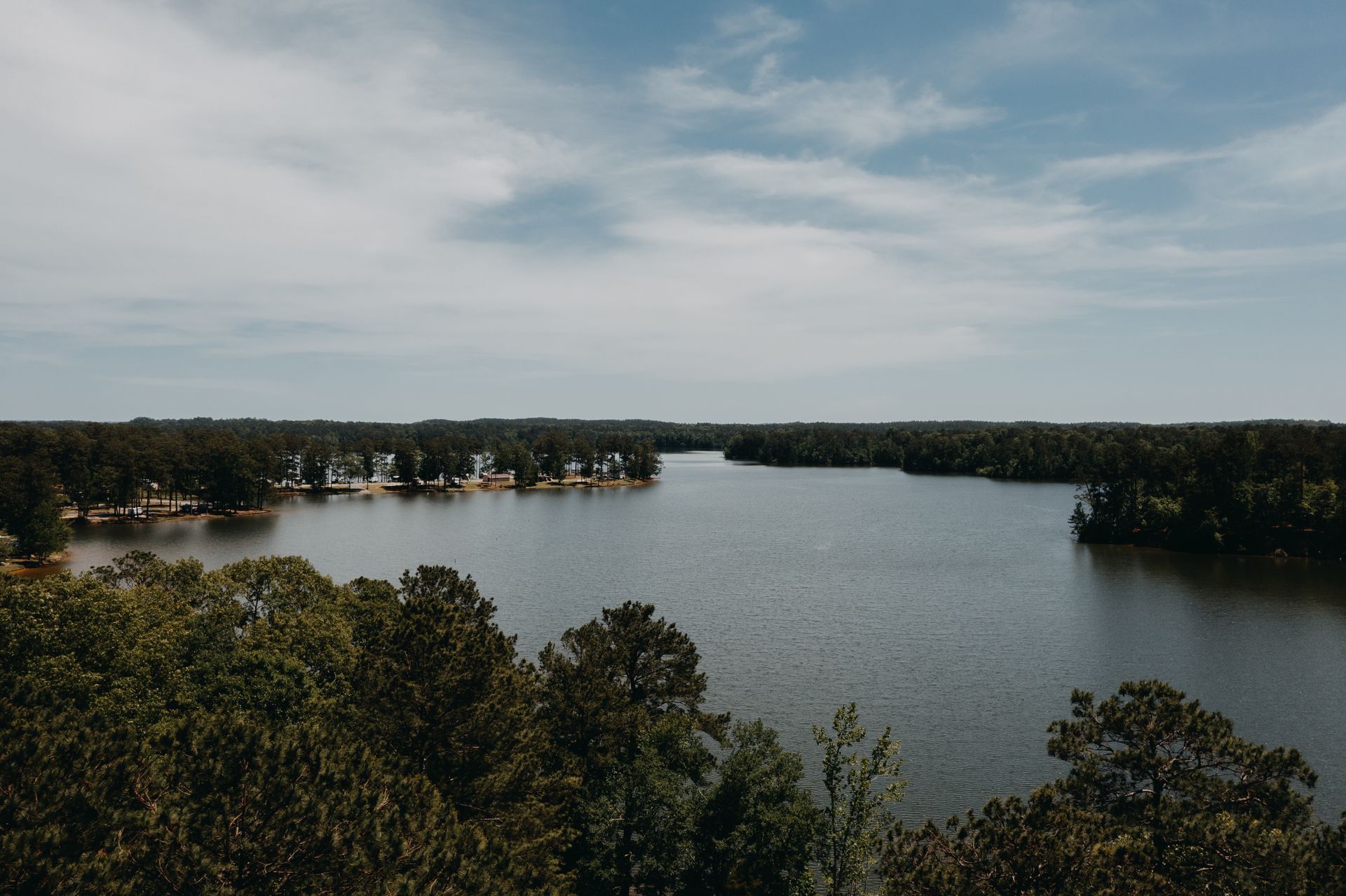 A wide view of a calm lake surrounded by trees under a cloudy sky.