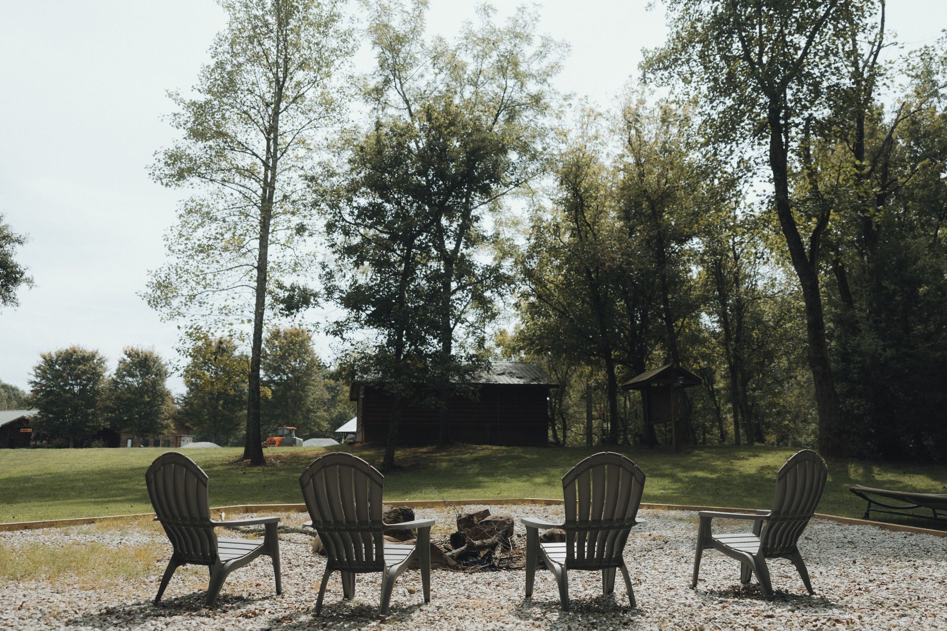 Four Adirondack chairs around a fire pit on a gravel patio, with trees and a cabin in the background.