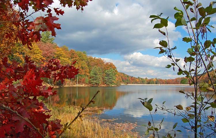 Autumn lake scene with vibrant red, orange, and yellow foliage reflecting in calm water under a partly cloudy sky.