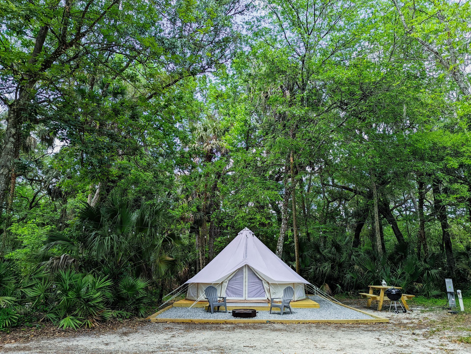 White canvas tent in a wooded campsite with two chairs, picnic table, and fire pit.