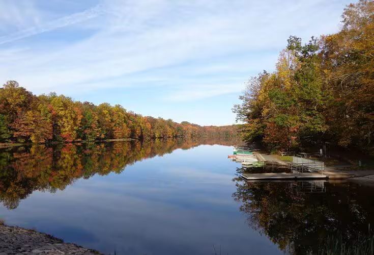 Calm lake reflecting autumn trees with red, orange, and yellow leaves under a blue sky.