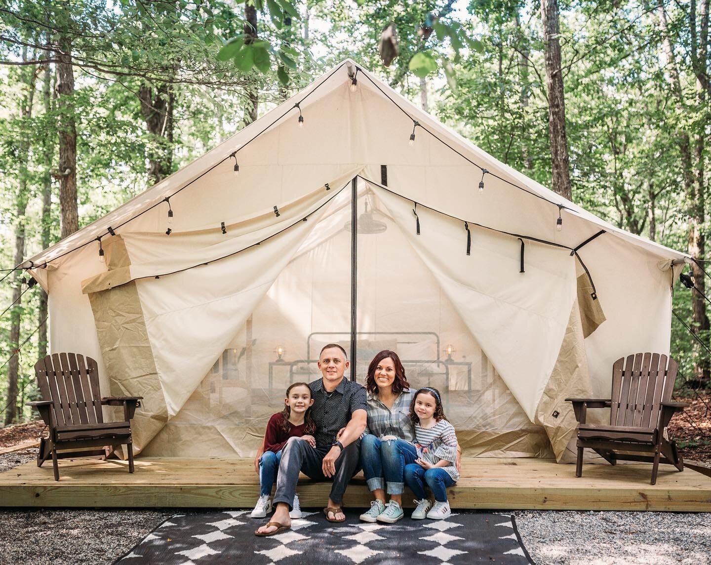 Family of four seated in front of a glamping tent in a wooded area; wearing casual clothes; smiling.