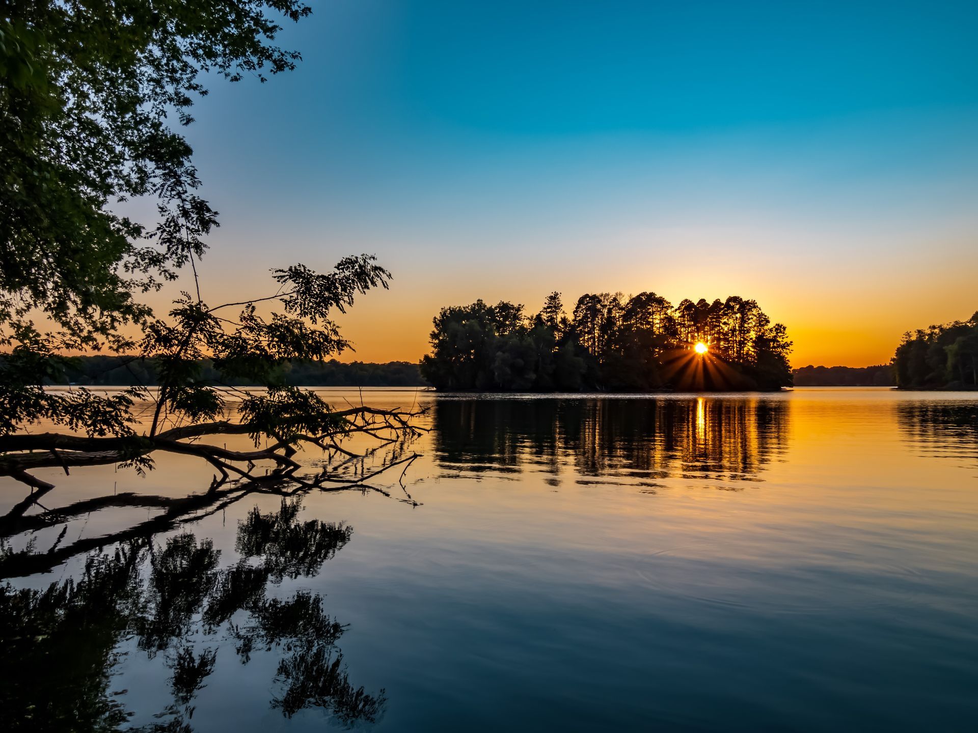 Sunset over calm lake, silhouetted island with trees, branches in foreground. Sky gradients of orange, blue.
