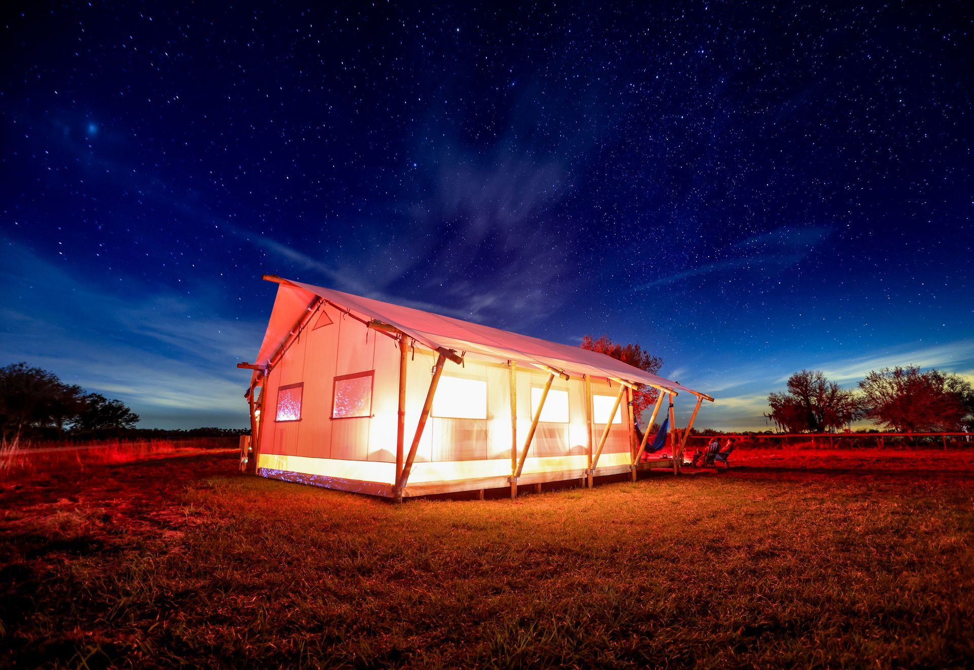 Lit-up canvas tent at night under a starry sky. Wooden frame, windows, and red ground lighting.