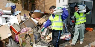 Two men are carrying bags of trash in front of a pile of trash.