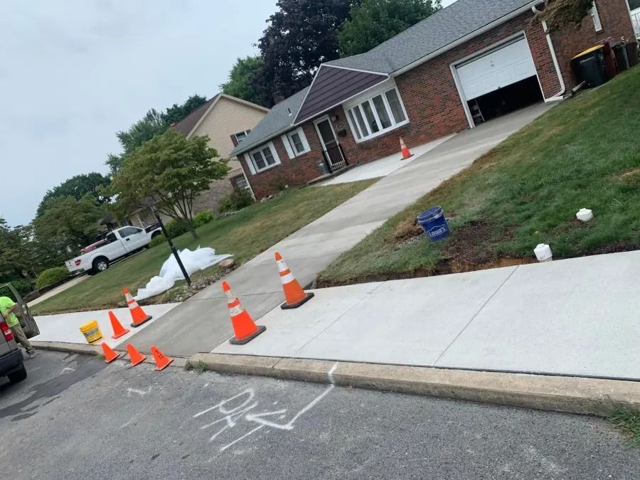 Cones blocking a suburban sidewalk beside a driveway and parked car on a sloped street.