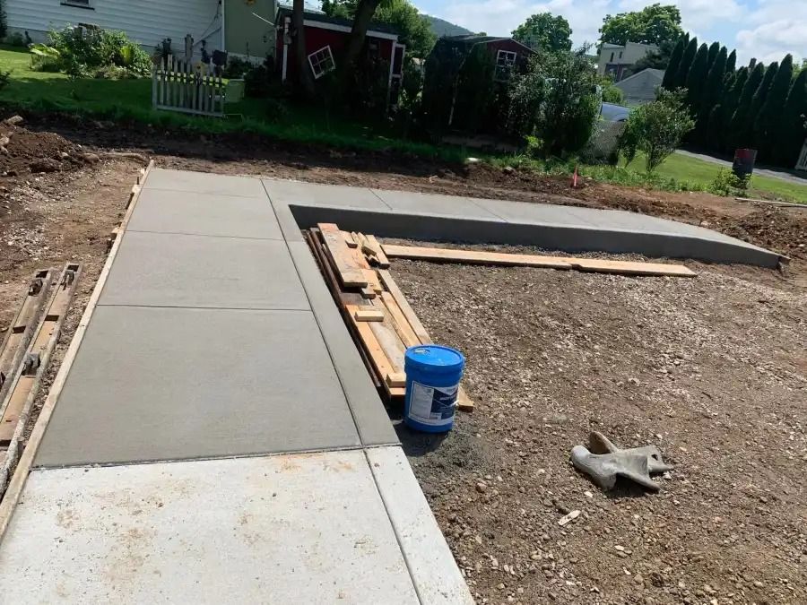 Concrete sidewalk under construction beside a gravel yard, with forms, lumber, and a blue bucket.