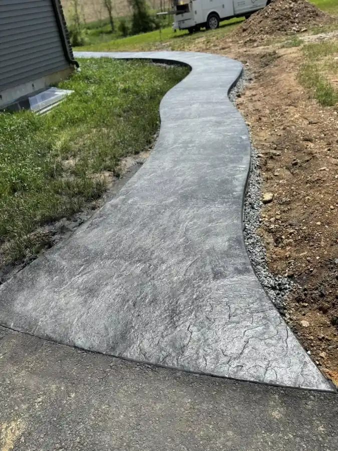 Curved concrete walkway under construction beside a house, bordered by grass and dirt.