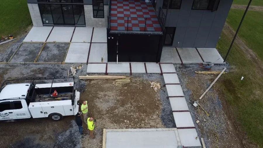 Aerial view of a construction site with a white utility truck, worker in a neon vest, and concrete walkway panels.