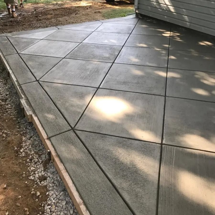 Concrete patio with geometric stamped sections beside a house, bordered by dirt and dappled sunlight.